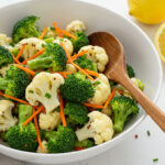 A vibrant broccoli cauliflower salad recipe in a white bowl, featuring fresh broccoli and cauliflower florets, shredded carrots, and a creamy dressing, topped with bacon bits and shredded cheese.