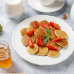 A plate of golden-brown mini pancakes stacked neatly, drizzled with maple syrup and topped with fresh blueberries and strawberries.